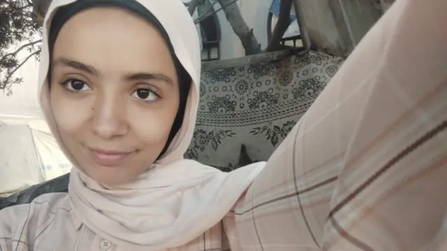 Eighteen-year-old Sanabel wearing a light pink hijab and dress, looks into the camera and smiles