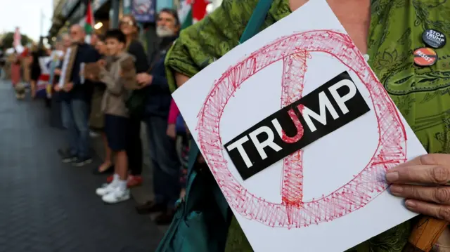 A demonstrator holds a white A4 sized portrait sign. It reads Trump on a black background in white lettering, with a hand drawn red coloured circle with a line going through it over it. behind the demonstrator is a  line of people