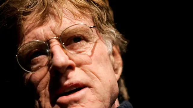 A headshot of Redford who looks up at an angle - against a dark background
