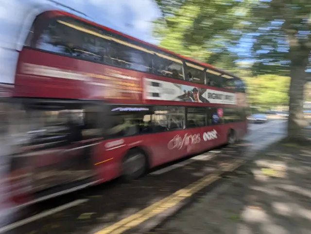 A blurry image of a red bus driving past the camera on a bright sunny day. The shutter speed has been adjusted to make it look like the bus is driving past quickly.