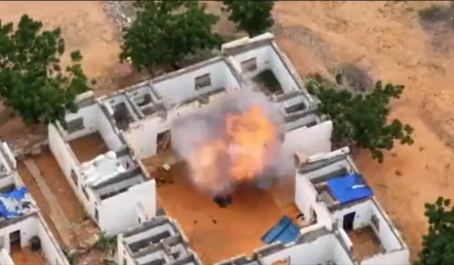 An explosion erupts in a ruined building in Sudan. The blast erupts at the back of a white building, surrounded by trees.