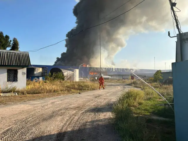 Smoke billows from a logistics centre in Ukraine. An emergency services officer can be seen walking toward the blaze
