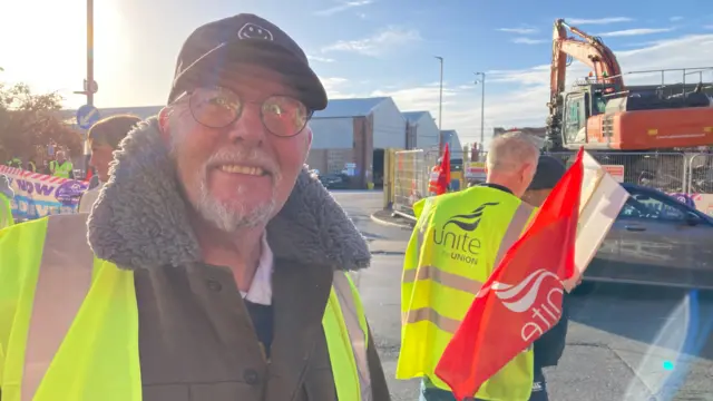 Simon Hendon wearing a yellow hi-vis vest, a fur lined jacket, round glasses and a black cap. He is standing at the picket line in front of the Lawrence Hill bus depot.