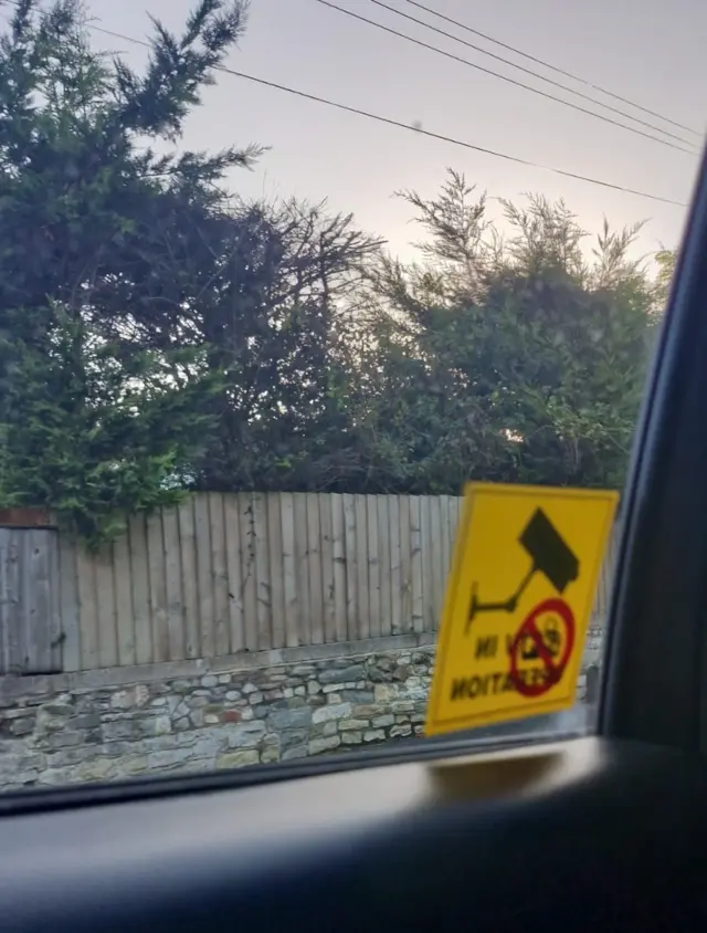 The view from the backseat window of a black taxi. There is a yellow sticker on the window, indicating there is video surveillance inside the car. Outside there is a wooden fence and some trees. It is dawn and the sky is grey and slightly pink.