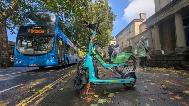 A blue-green Dott scooter and bike parked on the pavement in the foreground. In the background there is a blue number 2 bus driving along the road. There are lots of autumnal leaves on the ground and people walking past.