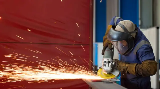 Sparks are seen as a man uses an angle grinder to cut and polish steel in a factory. he is wearing protective clothing and is standing on the right side of the picture, facing left.