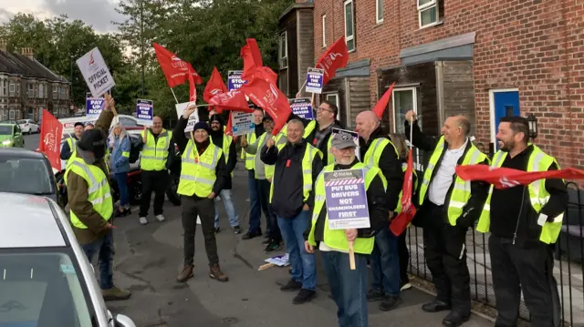 A large group of bus drivers standing beside the road wearing yellow hi-vis vests and holding up red flags and placards in protest. One says 'put drivers, not shareholders first'.