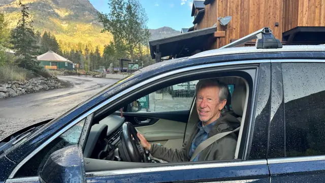 A man sits in his car and smiles as you see mountains in the background