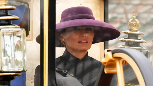 Melania Trump wearing a large rimmed purple hat, sat in a royal carriage in Windsor.