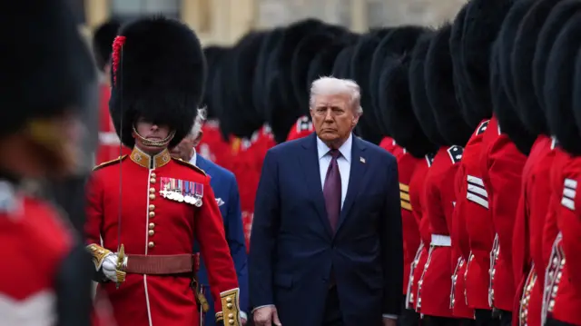 President Trump walks between rows the guard of honour dressed in red. He wears a navy suit.
