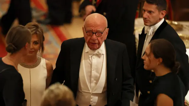 Rupert Murdoch stands at the centre of a group of people and appears to be mid-speaking