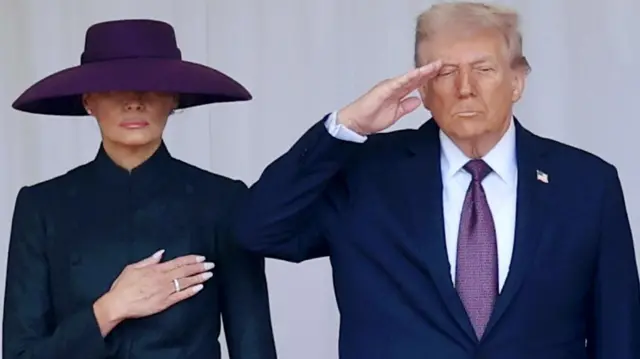 First Lady Melania Trump and President Donald Trump stand side by side with Melania holding her right hand to her chest and Trump saluting with his right hand