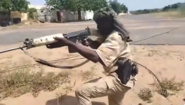 A video screenshot of a man in desert camouflage uniform aiming an assault rifle down a road, he is wearing a green turban