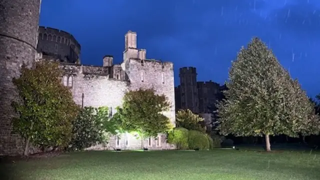 A wall of Windsor Castle looms behind trees under a dark blue sky.