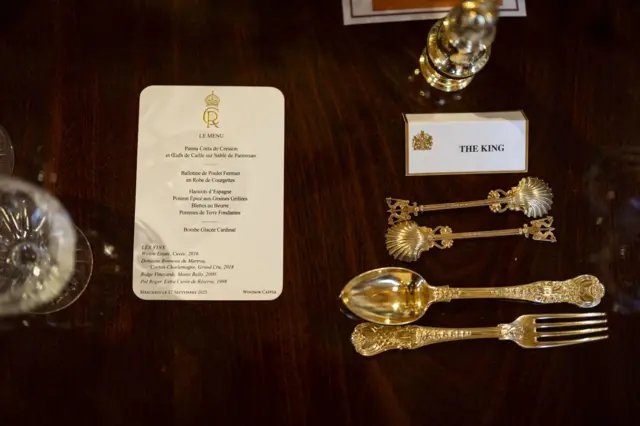 A close-up shot of the King's place laid at the banquet table. A name place reads 'The King'. The menu details the following dishes (written in French) - Starter: Hampshire watercress panna cotta with parmesan shortbread and quail egg salad  Main: Organic Norfolk chicken ballotine wrapped in courgettes with a thyme and savoury infused jus  Dessert: Vanilla ice cream bombe with Kentish raspberry sorbet interior with lightly poached Victoria plums
