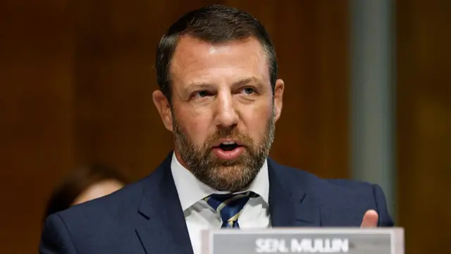 Sen Markwayne Mullen, a man in a navy suit with a salt and pepper beard and dark hair, sitting at a table with a name plate in front of him
