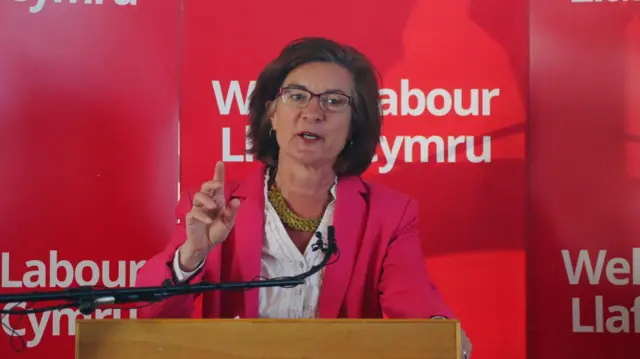 Eluned Morgan, wearing a pink jacket, speaks from a podium with a 'Welsh Labour' sign behind her.