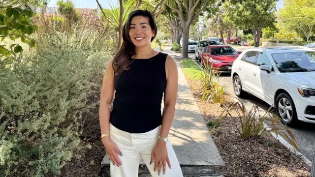 A woman wearing a black shirt and white pants poses on the sidewalk of a tree-lined residential street, next to a parked white car.
