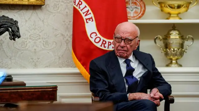 Murdoch sits on a chair with his hands clasped together. A Marine Corps flag is behind him