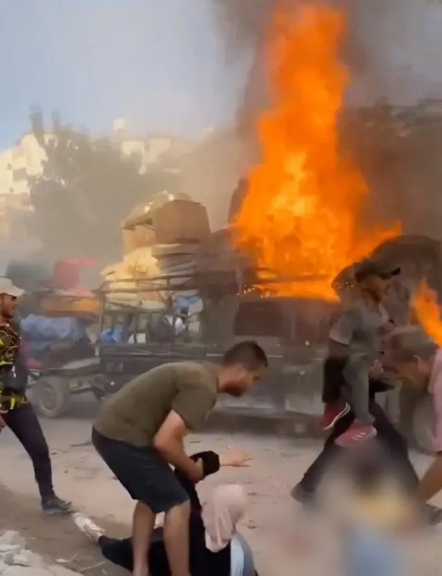 A pickup truck with a mattresses and other household belongings, the front of the vehicle has a huge flame burning from it. In the foreground is a man carrying a child in his arms, while another man is holding the arm of a woman in hijab on the ground.