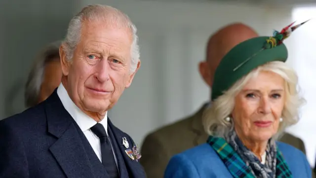 King Charles (L) wearing a navy suit and Queen Camilla (R) wearing a lighter shade of blue and a green fascinator