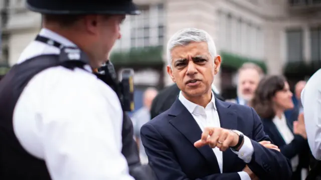 Sir Sadiq Khan, wearing a white shirt and a blue suit, speaks to a London police officer.