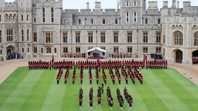 Wide shot of Donald Trump saluting troops from under a white canopy outside Windsor Castle, Melania Trump to his right, the King and Queen to his left