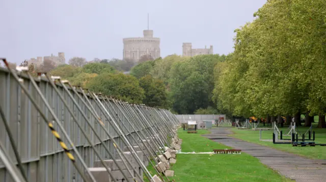 A long security fence stands on a grass lawn with trees and Windsor Castle in the background