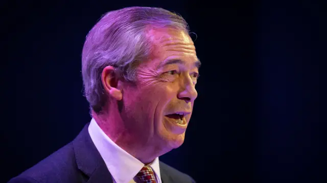 Nigel Farage speaking to the press wearing a suit with a red and silver checkered tie.