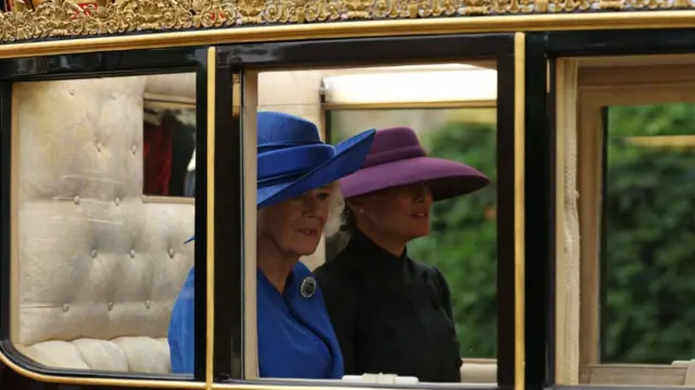 Queen Camilla (L) in blue hat and morning coat dress sits next to Melania Trump (R) in a purple hat and black dress