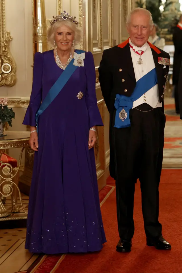 King Charles III on right in suit, Camilla on the left in a long aubergine coloured dress.