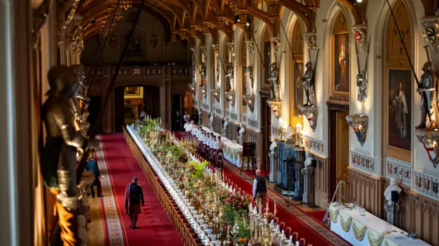 A landscape shot of the banquet table in St George's Hall. A number of places have been laid and the centre of the table is filled with flowers. Two men walk on either side of the table. A line of pipers can be seen on the right hand side of the table