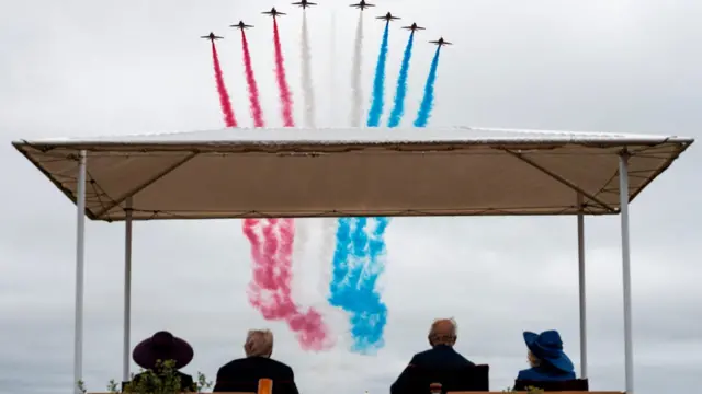 The king, queen president and first lady sit and watch the fly past ahead as red white and blue fills the sky.