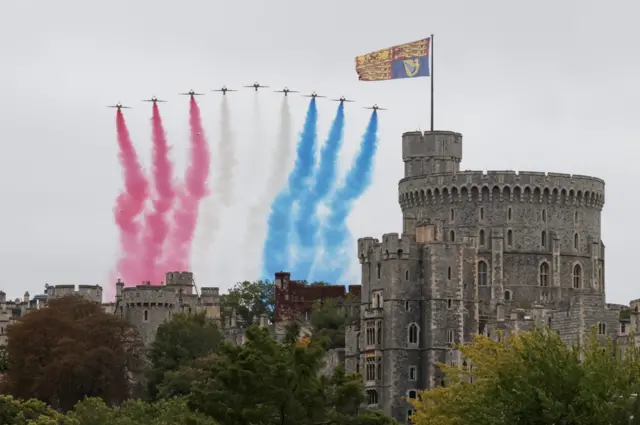 Red Arrows fly over Windsor Castle from an eastward approach. The flag of the King flies over the main tower of the castle