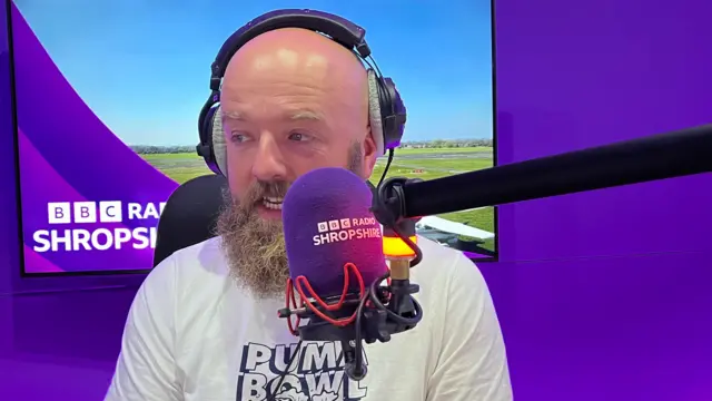 A man with a bald head and light brown beard wearing headphones with a BBC Radio Shropshire-branded TV behind him