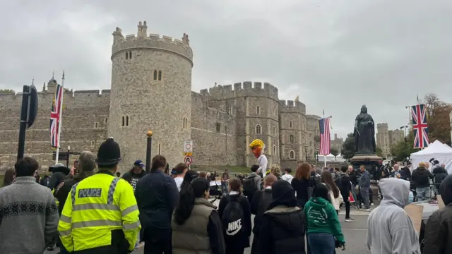 Crowd stands in foreground, looking towards Windsor Castle in the background. In the middle is a sculpture of Donald Trump raised aloft - he's made to look like a baby in a white babygrow.