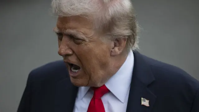 A close up photo of Donald Trump wearing a dark blue suit and a red tie with US flag pin on his collar