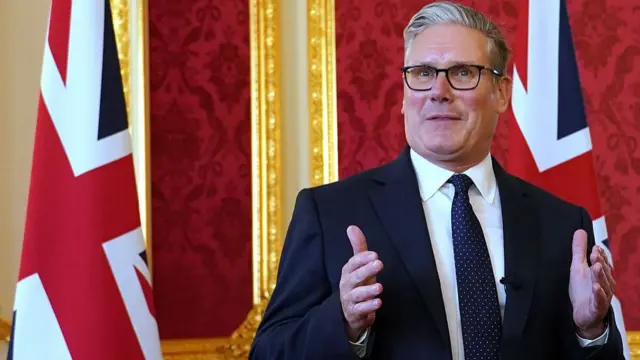Close up of Prime Minister Sir Keir Starmer in a ornate room with walls covered in red brocade, two Union flags on poles behind him
