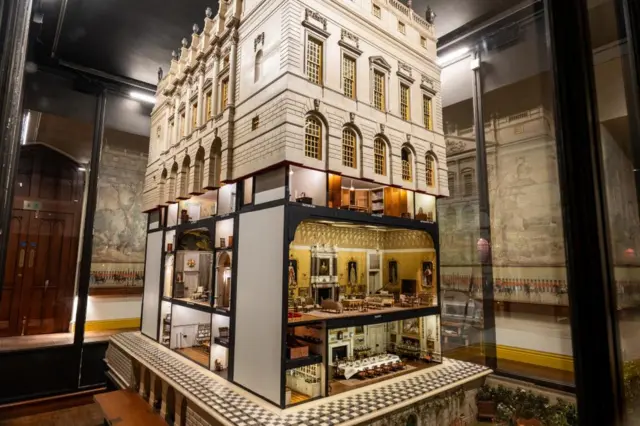The Queen Mary's Dolls' House - a towering miniature of while brick walls and golden windows, with the bottom two floors open for viewing of a golden drawing room and lavish dining room