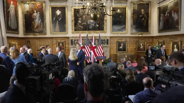 Media setting up cameras in a grand room with US and UK flags, as people mill around