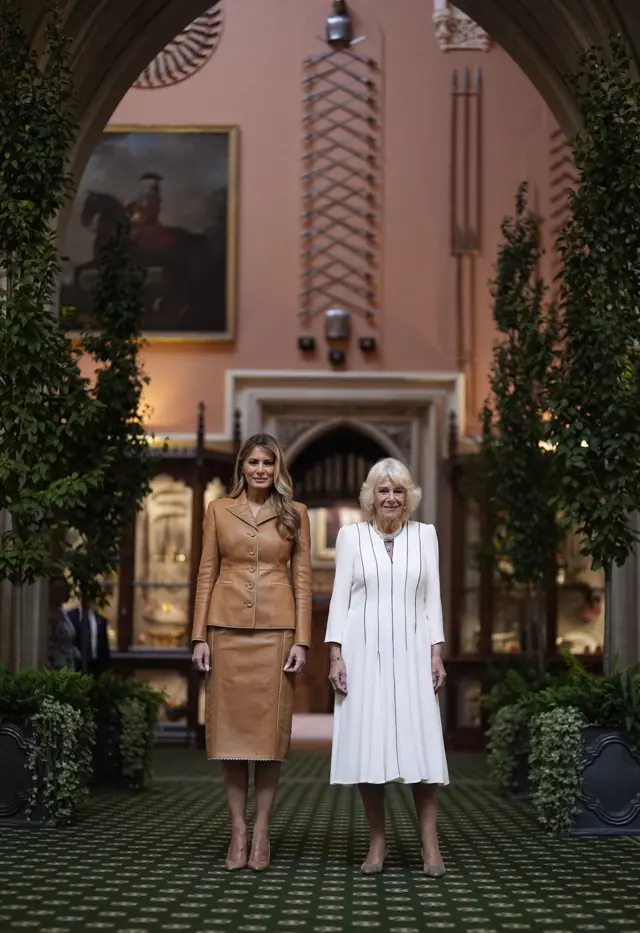 Melania Trump (L) in a light brown leather skirt suit stands next to Queen Camilla (R) in a white morning cocktail dress