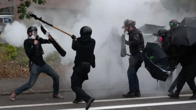 Around 5 Antifa and far-right protesters fighting. They are wearing gas masks and other protective gear like body armour, helmets, ski goggles and shields. One man is swinging a large stick at another who is holding a shield up, while someone further back fires some kind of pepper spray or tear gas. tear gas clouds are visitibe