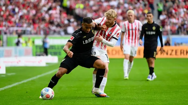 Stuttgart winger Jamie Leweling wearing a black kit shielding the ball from a Freiburg player in red and white