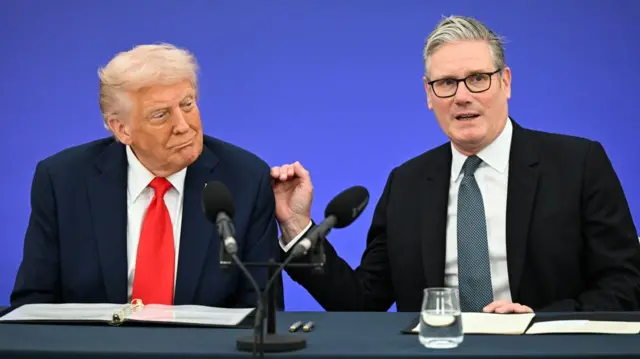Donald Trump (L) in black suit and red tie sits next to Keir Starmer (R) in a black suit and blue tie. Starmer is touching Trump's left shoulder with his hand