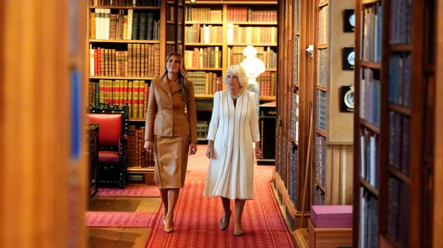 Queen Camilla on the right and First Lady Melania Trump on the left walking through the royal library in formal attire.