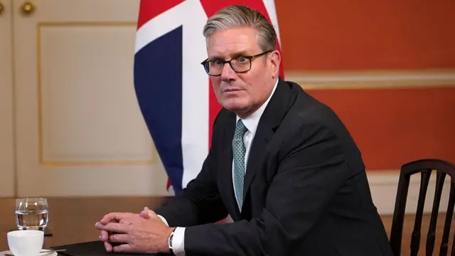 Close up of Keir Starmer looking pensive while sitting down on a wooden chair, his hands leaning on a desk. There's a coffee cup and a glass of water in front of him