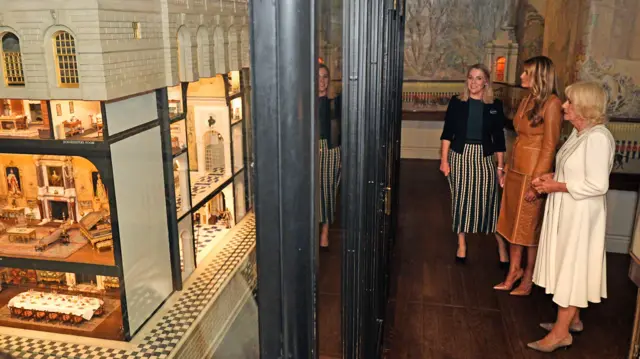 On the left is Queen Mary's Dolls' House on display. To the left are a guide outlining the contents to Melania Trump (L) and Queen Camilla (L)