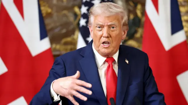 Donald Trump gesticulating as he answers questions at press conference. He's in a dark blue suit and red tie, two Union Jacks and a US flag on poles behind him