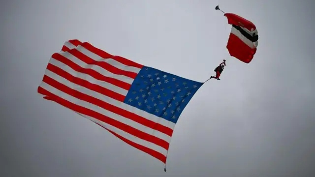 A paraglider with the US flag behind them glides through an overcast sky