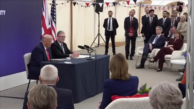 Trump and Starmer sitting at a table at the earlier statement, and the audience of business leaders and other politicians can be seen
